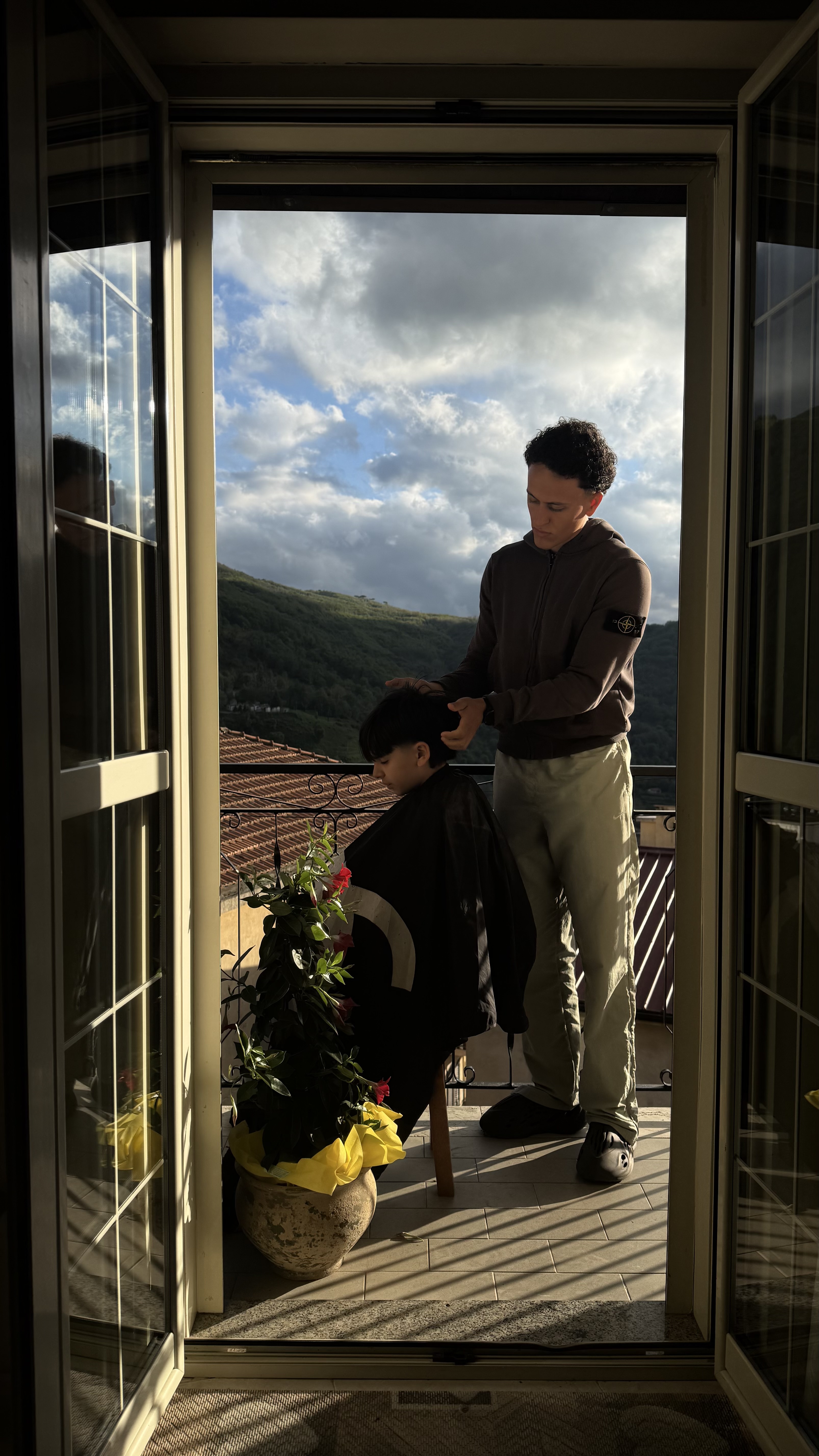 Mehdi coiffe un client sur un balcon, entre lumière naturelle et paysage ouvert.